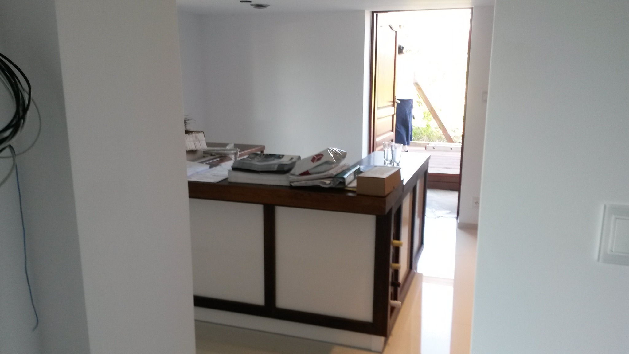 Biała wyspa kuchenna z ciemnobrązowym blatem, na którym leżą papiery, kalkulator i szklanki, widoczna z boku w jasnym wnętrzu z otwartymi drzwiami na zewnątrz.