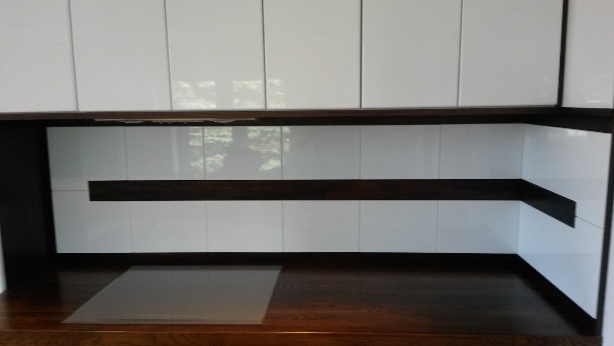 Nowoczesna kuchnia z białymi, lakierowanymi frontami szafek i ciemnobrązowym blatem z drewna, widoczny fragment płyty grzewczej zabezpieczonej folią.