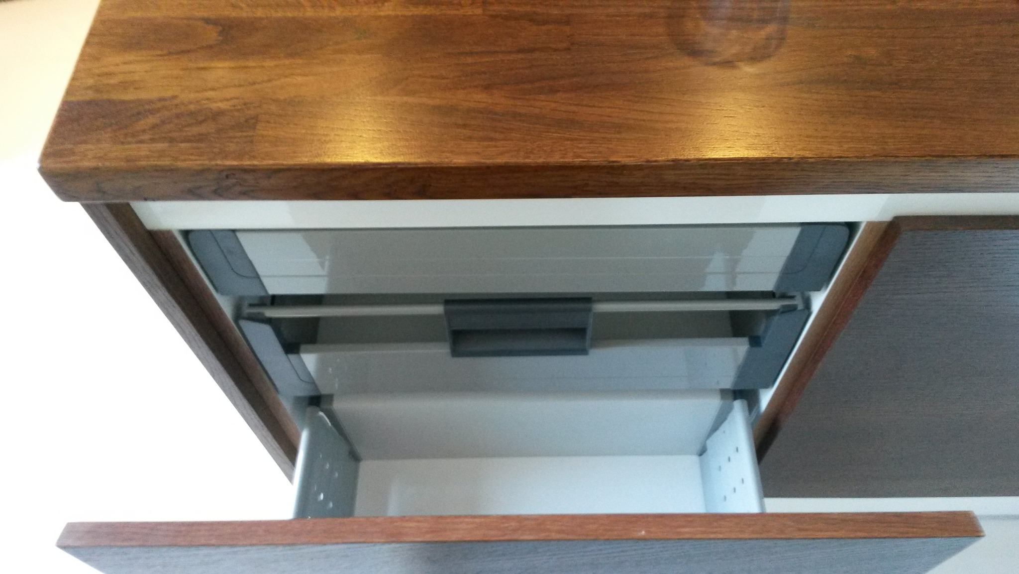 Otwarta szuflada z drewnianym frontem i metalowymi prowadnicami, zamontowana pod blatem z ciemnego drewna, widok z dołu.