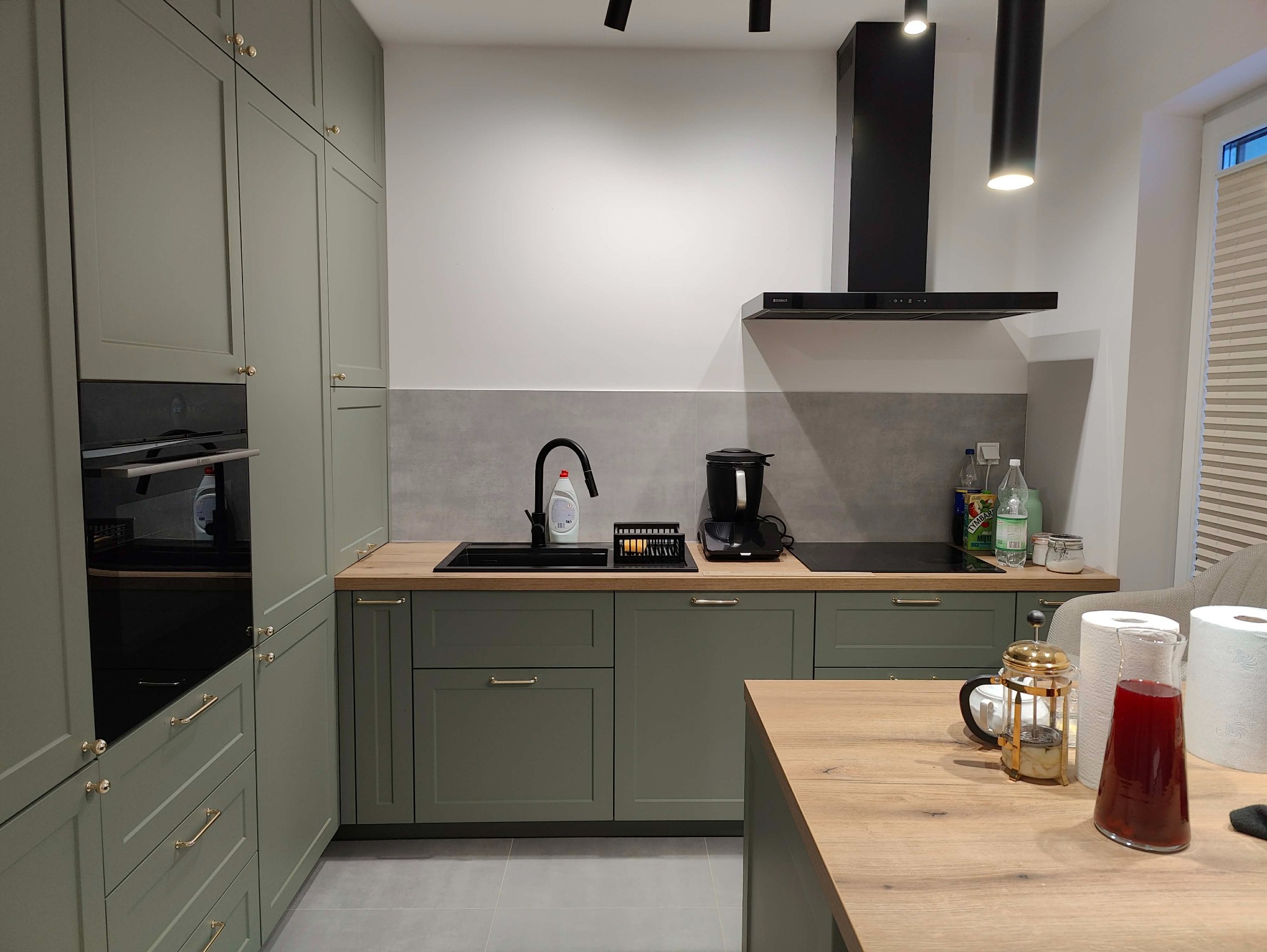 Nowoczesna kuchnia w odcieniach zieleni z drewnianym blatem, czarną armaturą i oświetleniem punktowym. Widoczny ekspres do kawy i akcesoria kuchenne.