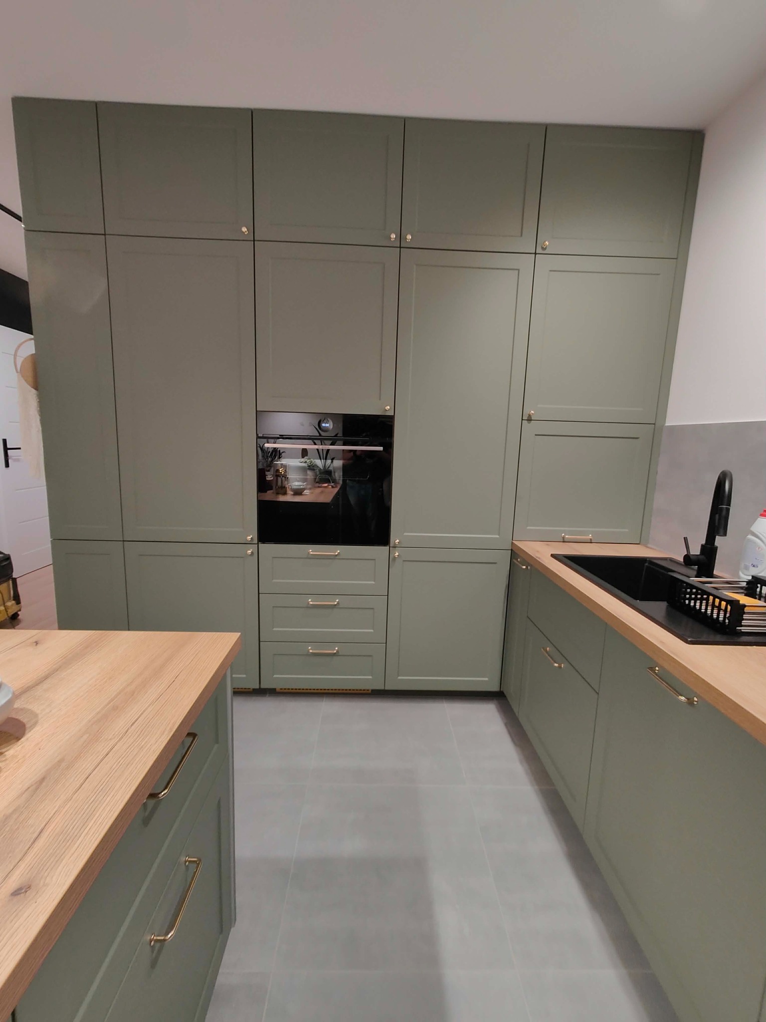 Kuchenna zabudowa w odcieniach zieleni z drewnianym blatem, złote uchwyty, piekarnik w zabudowie. Minimalistyczny design, szare płytki podłogowe.