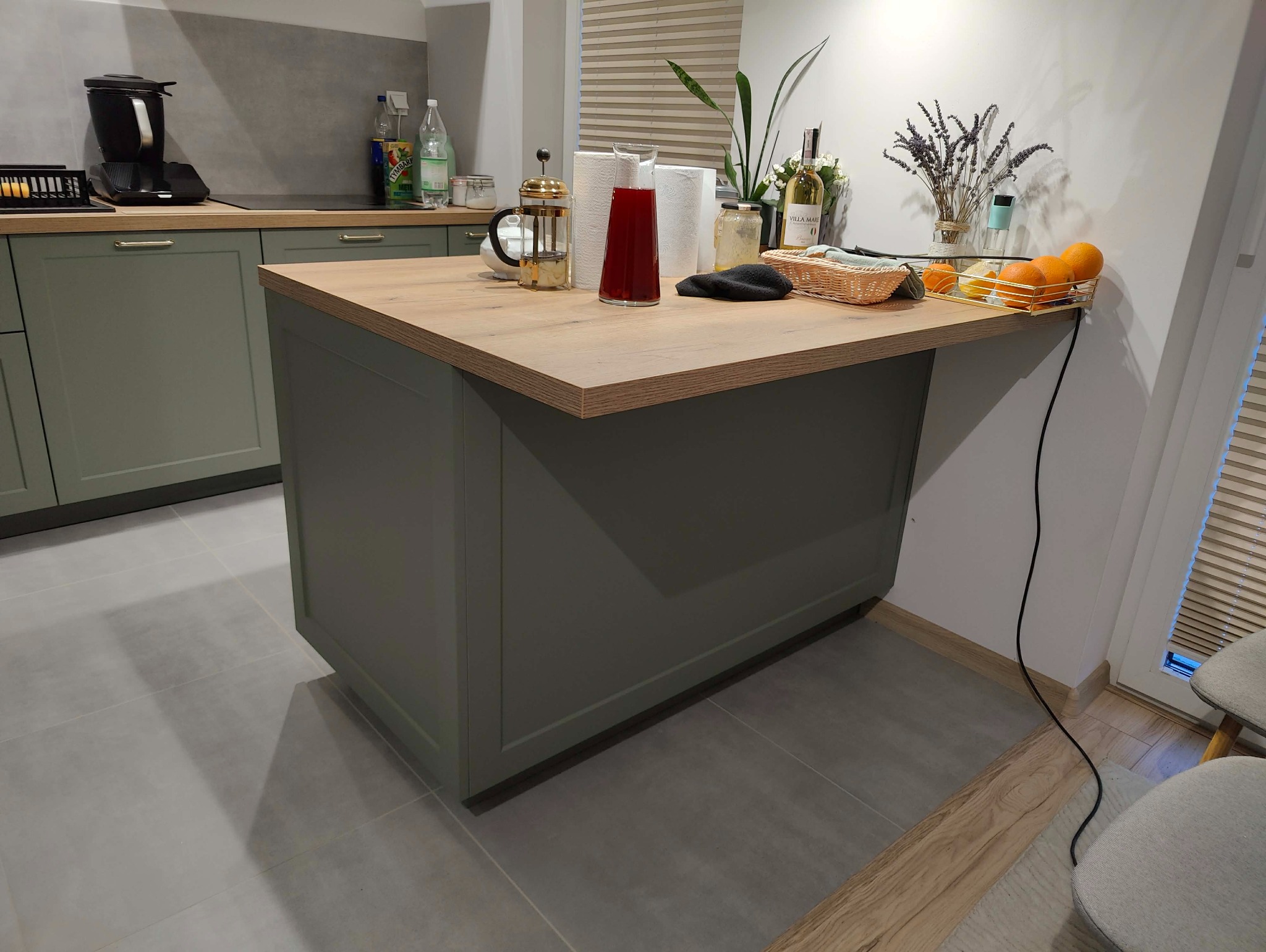 Nowoczesna kuchnia z blatem drewnianym i zielonymi szafkami. Na blacie owoce, dzbanek z napojem i prasa francuska. Minimalistyczny design, jasne wnętrze.