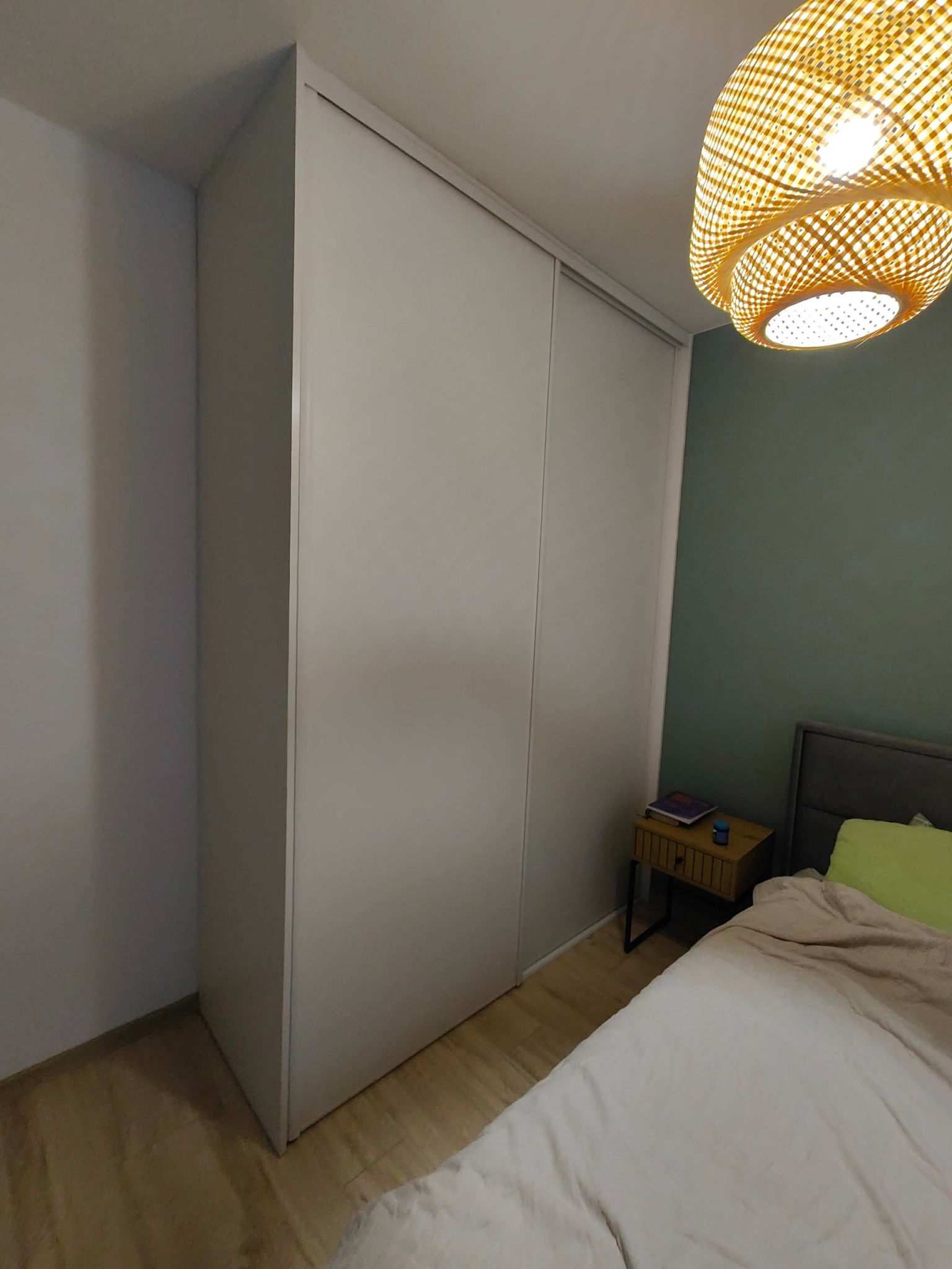 Minimalistyczna szafa wnękowa z przesuwnymi drzwiami w jasnym odcieniu szarości, wkomponowana w aranżację sypialni z drewnianą podłogą i lampą z abażurem z wikliny.