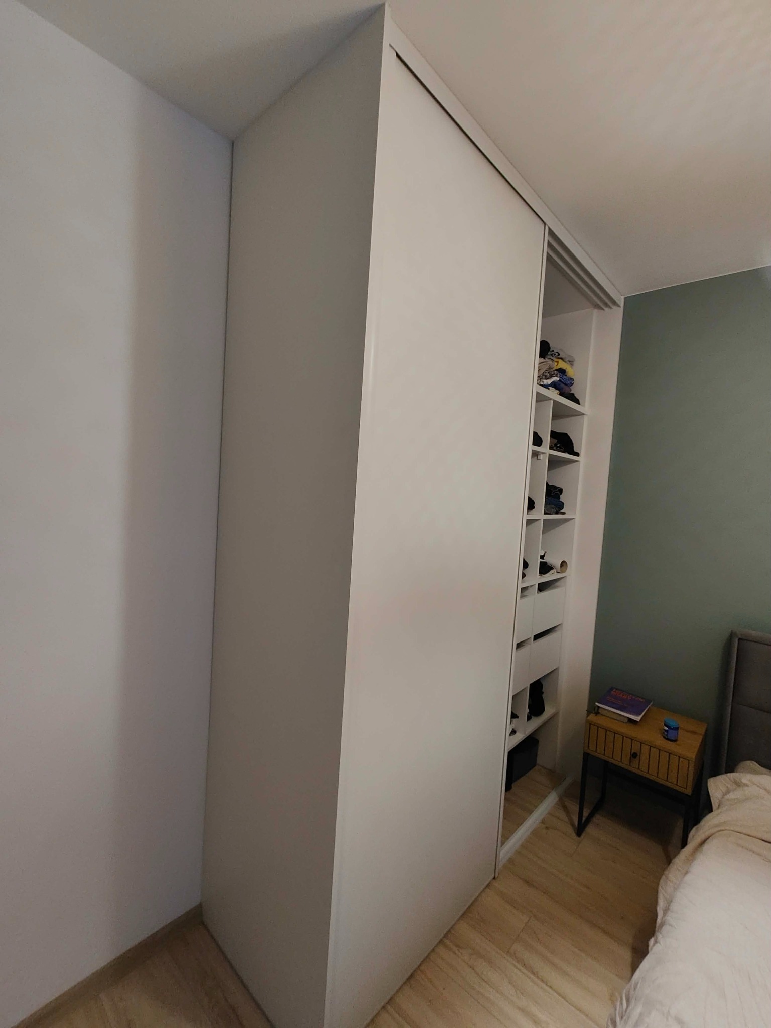 Biała szafa wnękowa z przesuwnymi drzwiami, częściowo otwarta, ukazująca półki z ubraniami. Widoczna szafka nocna i fragment łóżka. Minimalistyczny design.