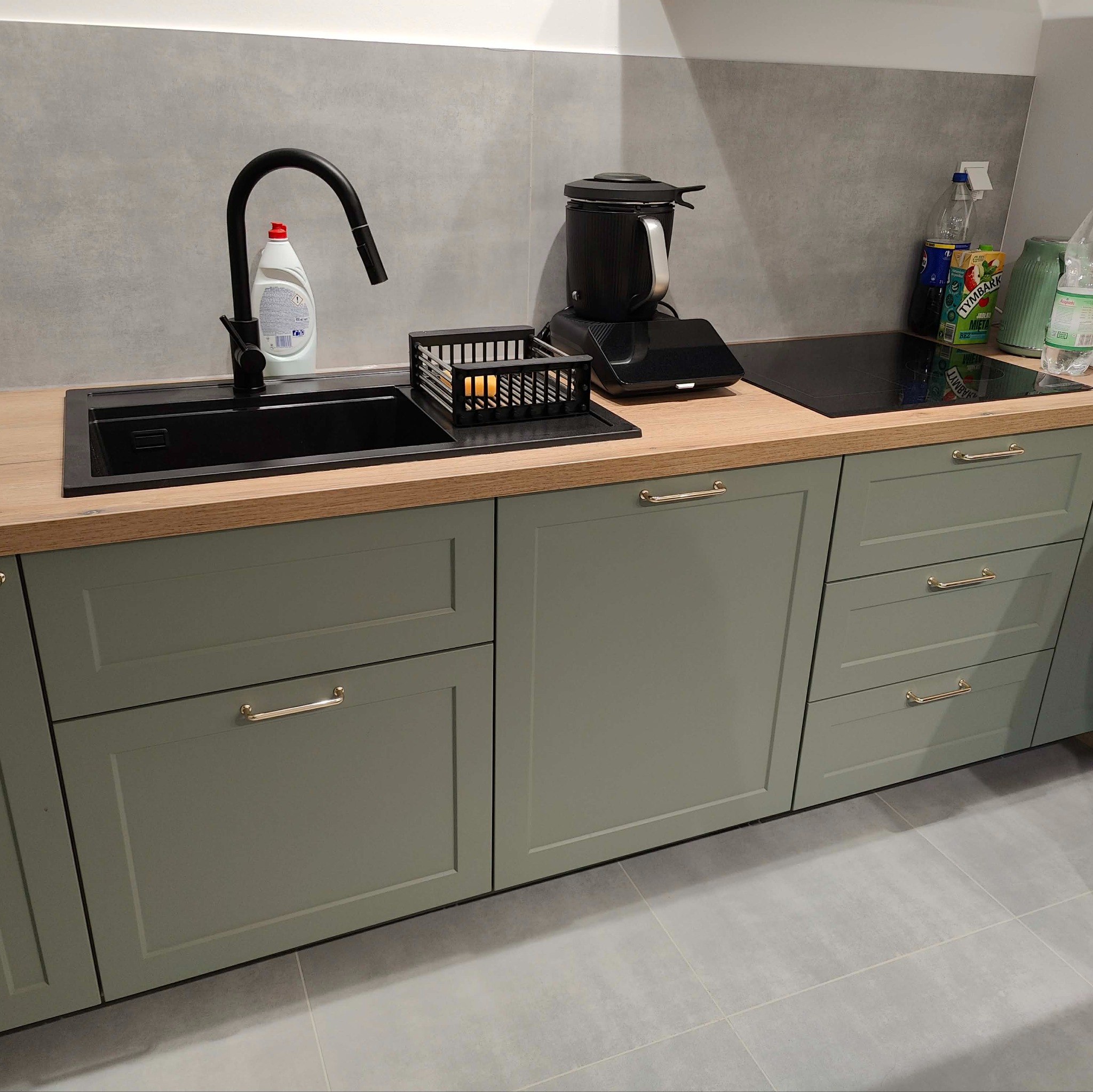 Fragment kuchni z drewnianym blatem, czarnym zlewem i baterią. Fronty szafek w odcieniach zieleni z delikatnymi złotymi uchwytami. Na blacie akcesoria kuchenne.