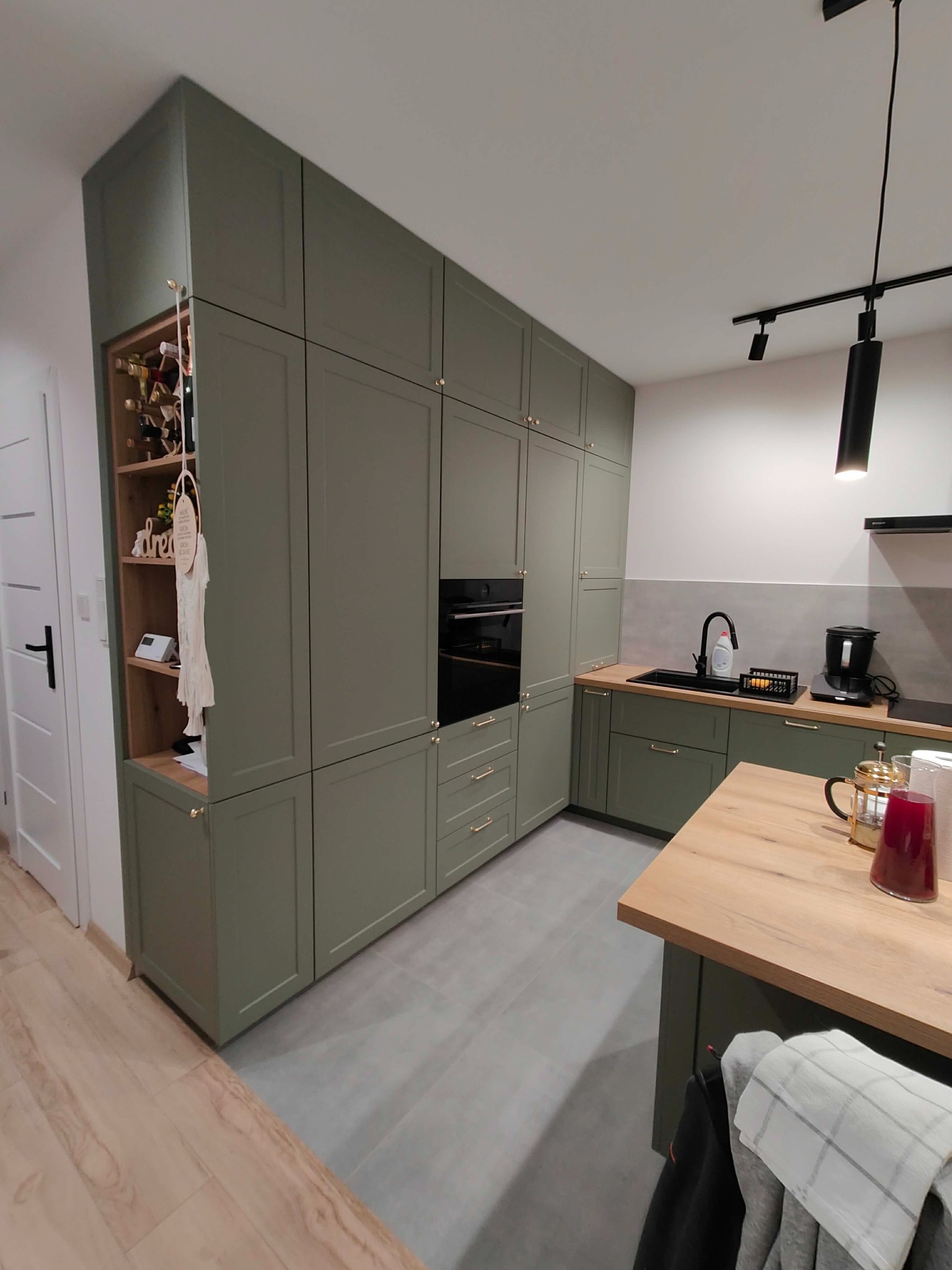 Kuchenna zabudowa w odcieniach zieleni z drewnianym blatem i otwartą półką na wino. Minimalistyczne oświetlenie punktowe. Nowoczesny design.