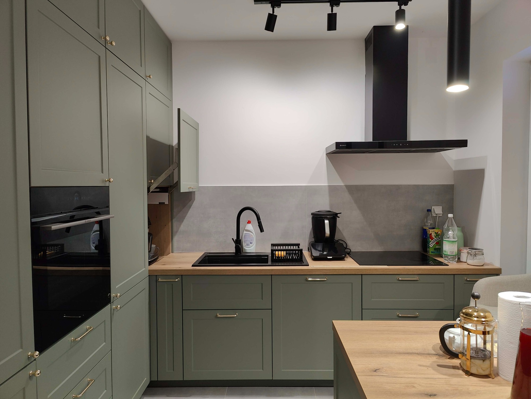 Kuchenna zabudowa w odcieniach zieleni z drewnianym blatem, czarnym zlewem i baterią. Nad blatem czarny okap i oświetlenie punktowe. Na blacie akcesoria kuchenne.