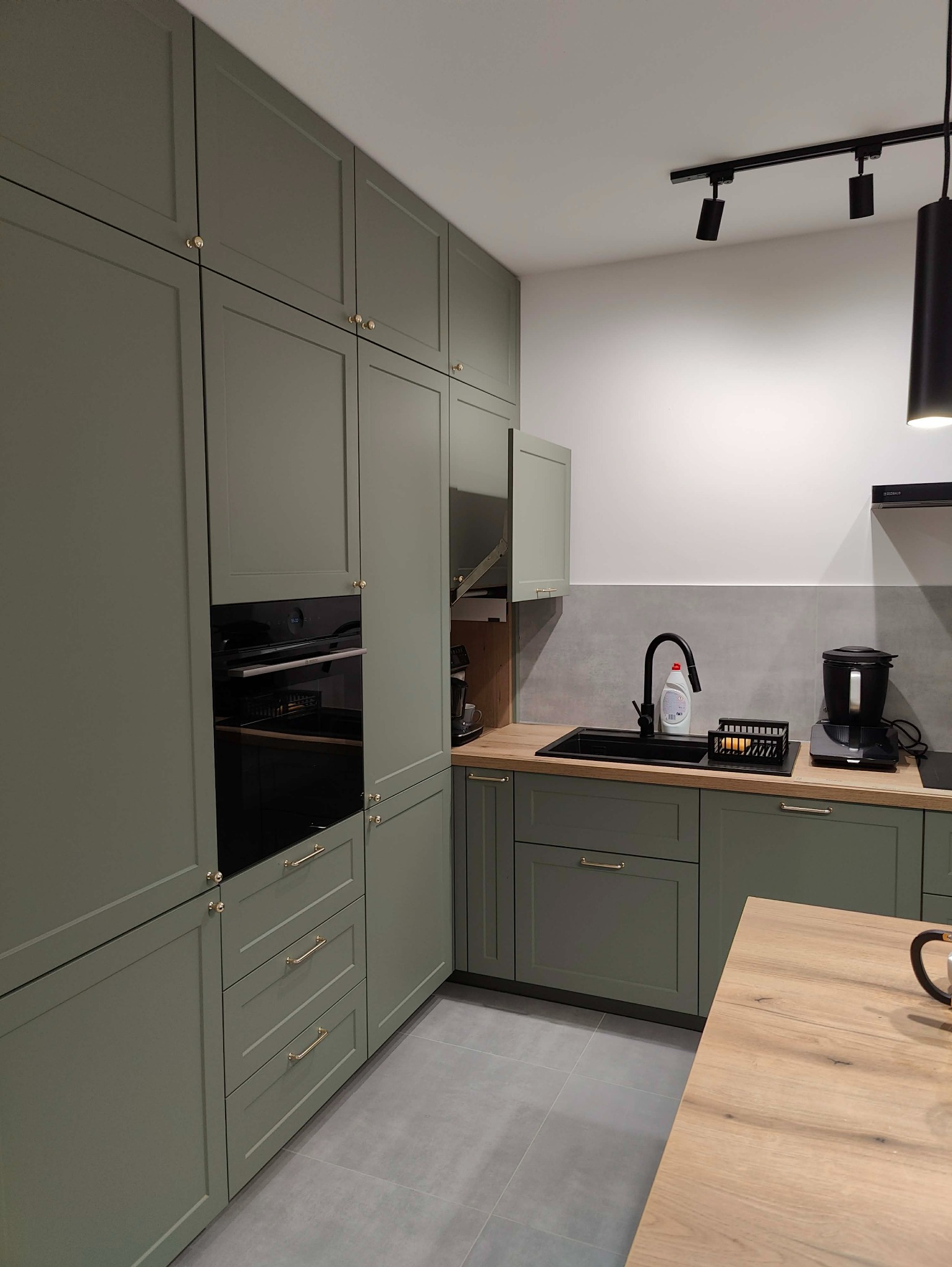 Kuchenna zabudowa w odcieniach zieleni z złotymi uchwytami, czarnym zlewem i sprzętem AGD. Widoczne oświetlenie punktowe i fragment drewnianego blatu.