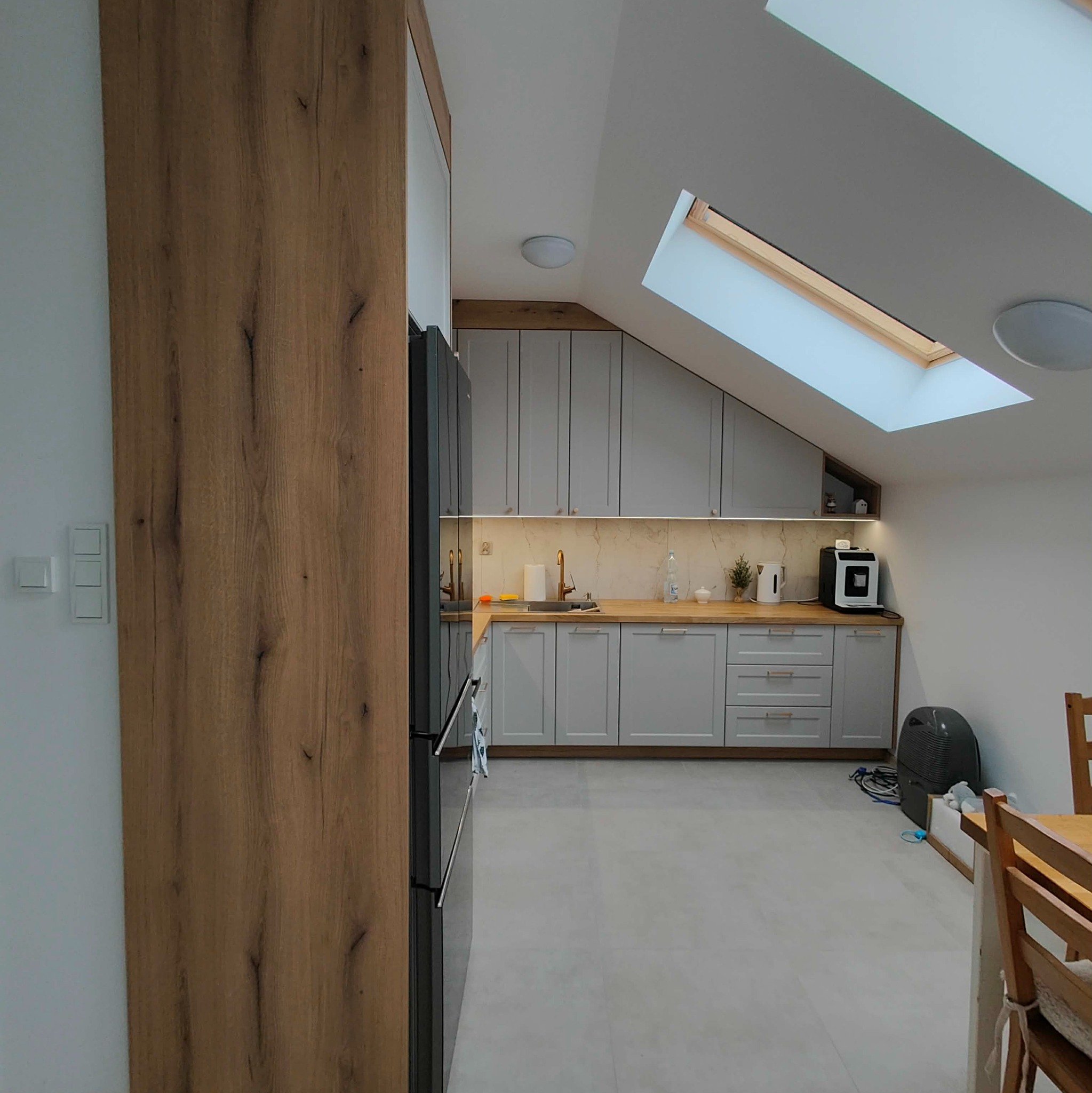 Jasna kuchnia na poddaszu ze skośnym oknem dachowym. Szare szafki, drewniany blat i wysoka zabudowa z forniru. Minimalistyczny styl, oświetlenie LED pod szafkami.