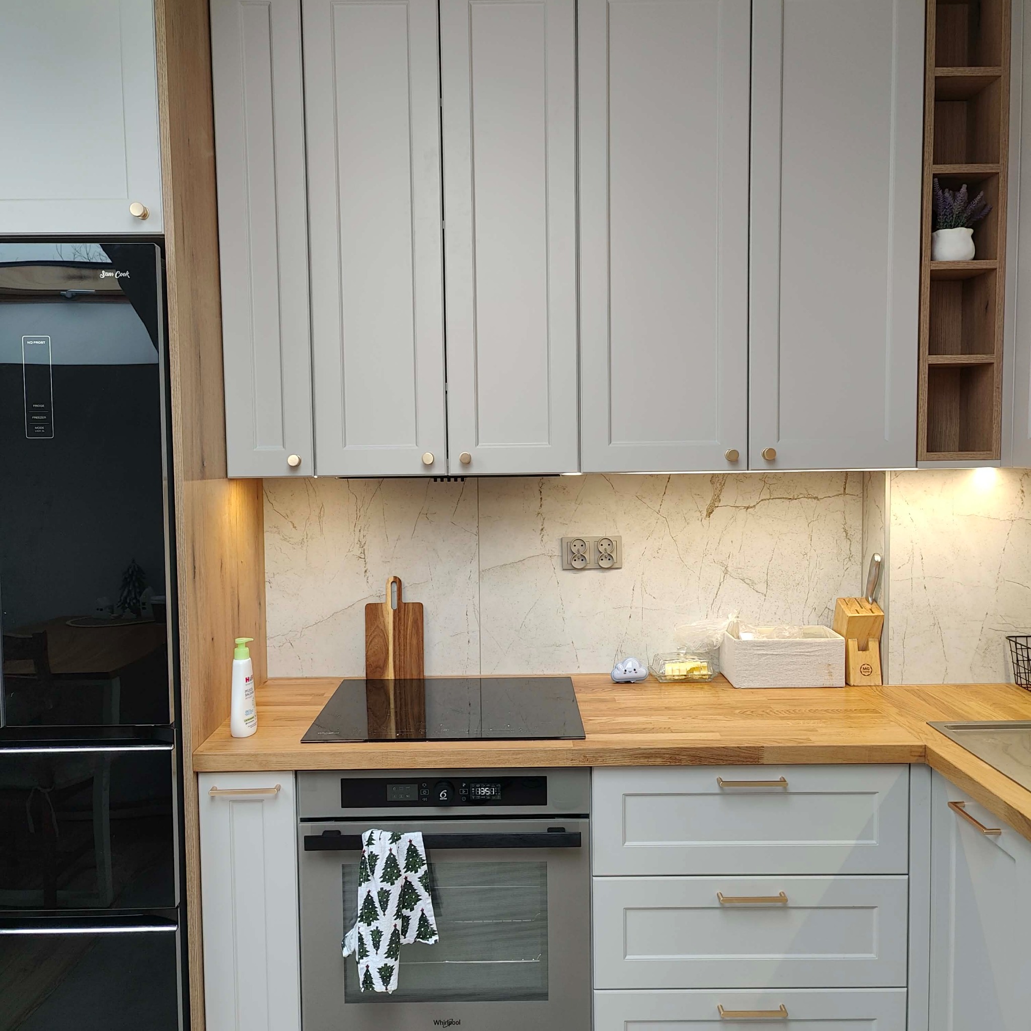 Jasnoszara kuchnia z drewnianym blatem i złotymi uchwytami. Na blacie płyta indukcyjna, deska do krojenia i akcesoria kuchenne. Widoczny fragment lodówki i piekarnika.