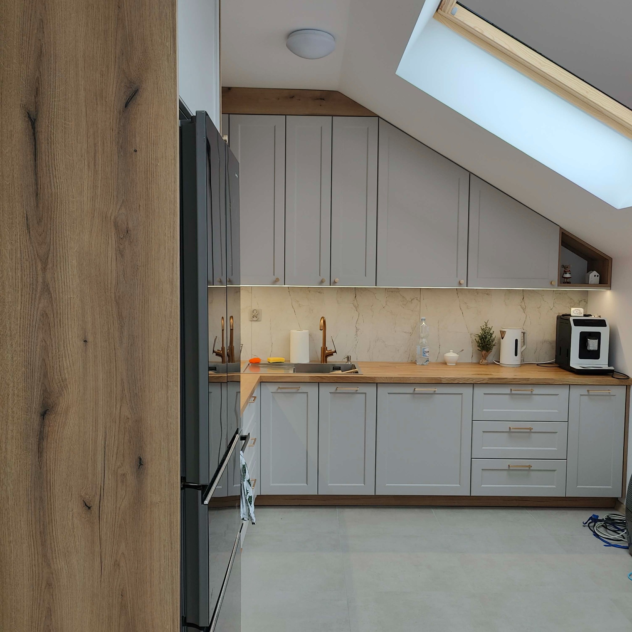 Jasna kuchnia z drewnianym blatem i oknem dachowym. Szafki w zabudowie pod skosem, złote akcenty. Nowoczesny design z elementami naturalnego drewna.