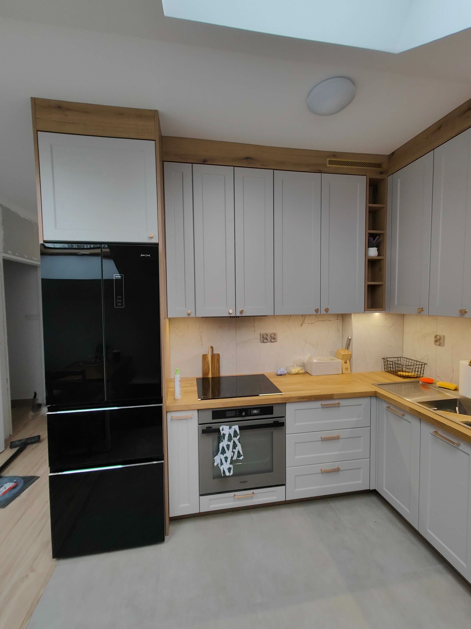 Jasnoszara kuchnia z drewnianymi blatami i czarną lodówką side-by-side, oświetlona lampą sufitową i światłem z okna dachowego.