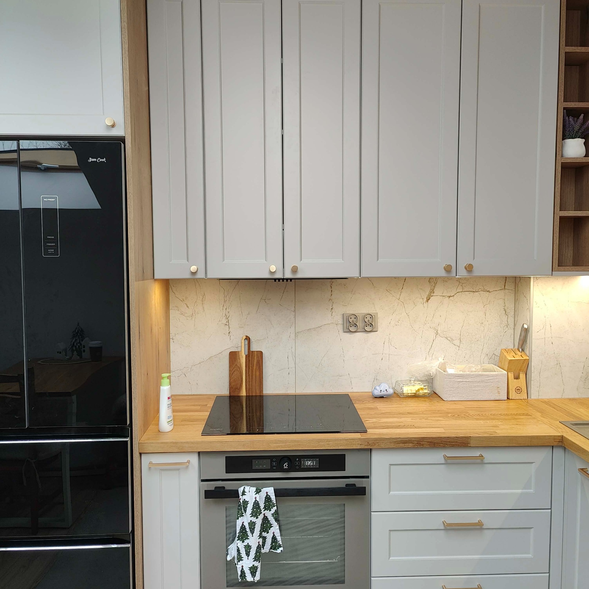 Jasnoszara kuchnia z drewnianym blatem, czarną płytą indukcyjną i piekarnikiem. Na blacie deska do krojenia i akcesoria kuchenne. Widoczna lodówka z czarnym frontem.