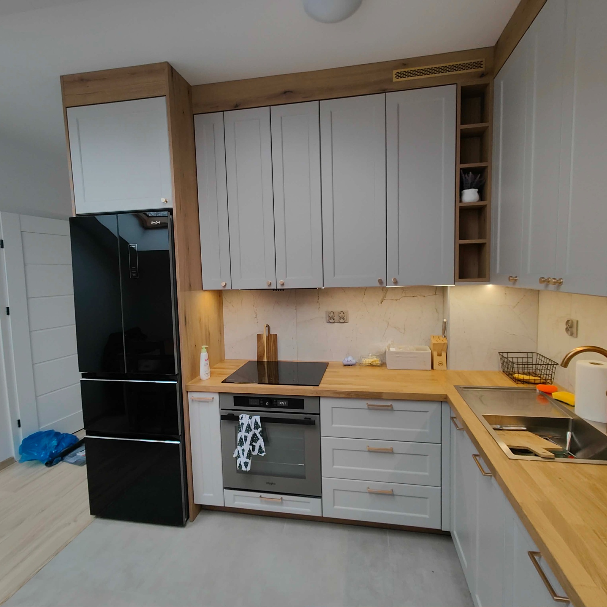 Jasna kuchnia z drewnianym blatem i szarymi szafkami, czarna lodówka side-by-side. Uwagę przyciąga drewniana zabudowa górnych szafek i półki na przyprawy.