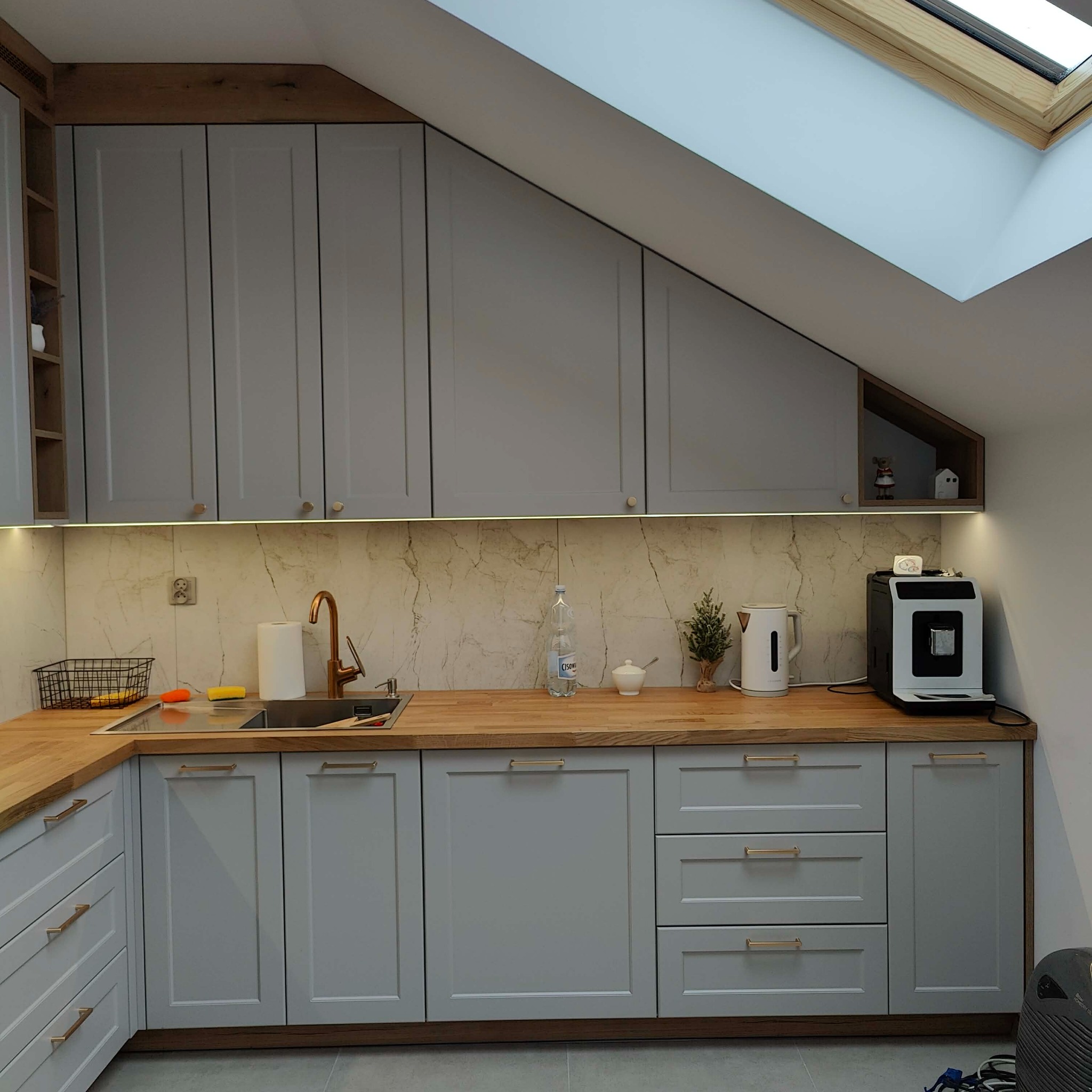 Szara kuchnia na wymiar z drewnianym blatem pod skosem dachu i oknem dachowym. Uwagę przykuwa złota bateria i marmurowy wzór na ścianie.