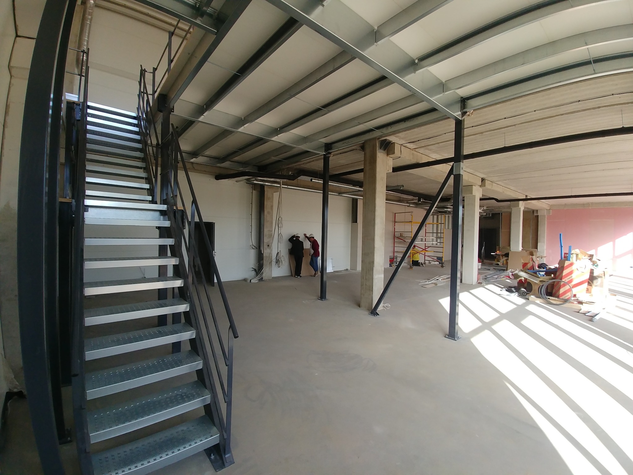 Wnętrze hali magazynowej w trakcie budowy lub adaptacji, widoczne metalowe schody, betonowe słupy, instalacje pod sufitem oraz osoby w oddali.