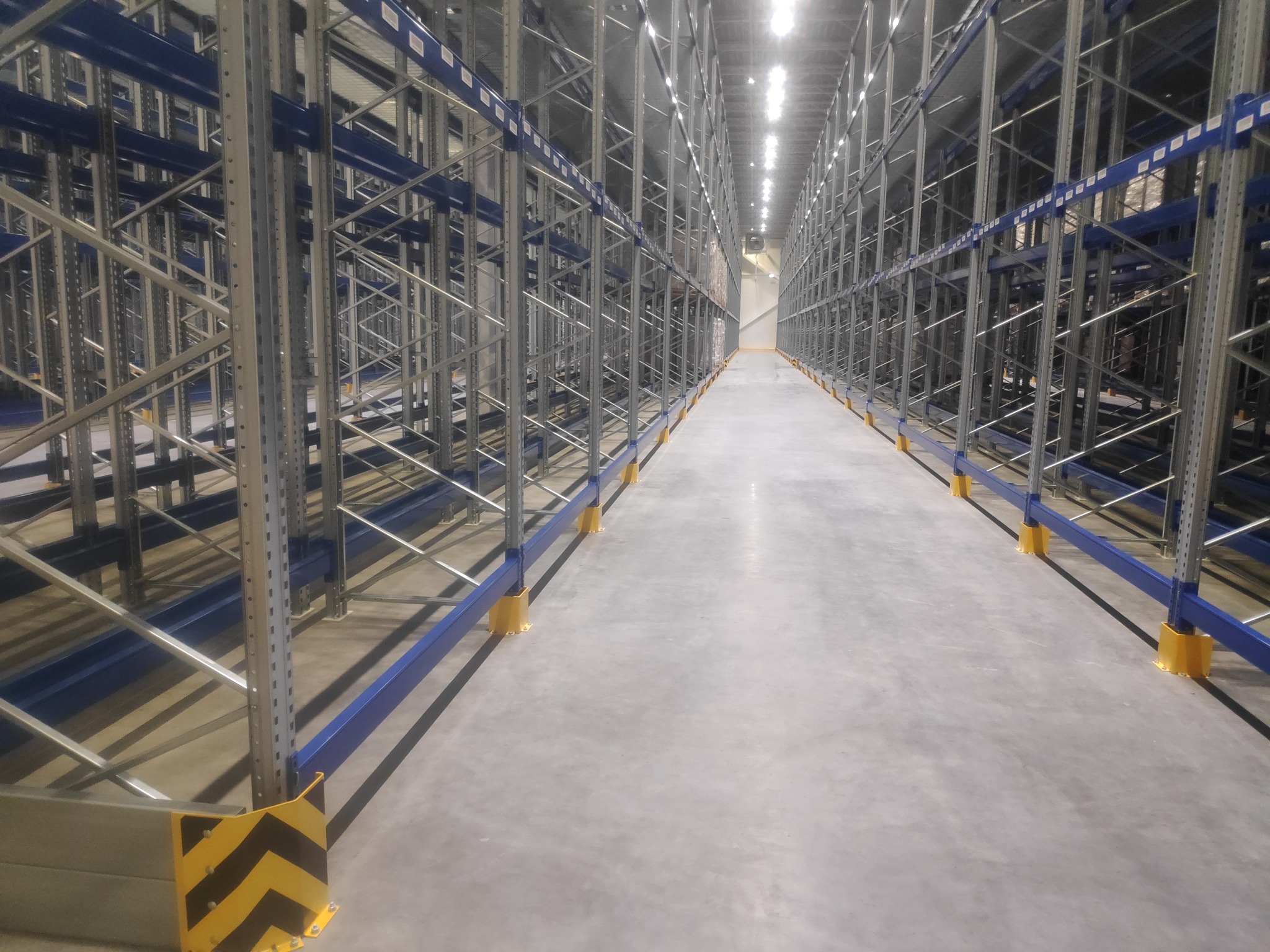 Długi korytarz między rzędami metalowych regałów magazynowych z niebieskimi poprzeczkami i żółtymi osłonami słupów, widok z perspektywy podłogi w głąb magazynu.