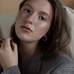 Portret kobiety z delikatnym makijażem, w tweedowej marynarce, pozującej w subtelnym oświetleniu, z widocznymi złotymi kolczykami.