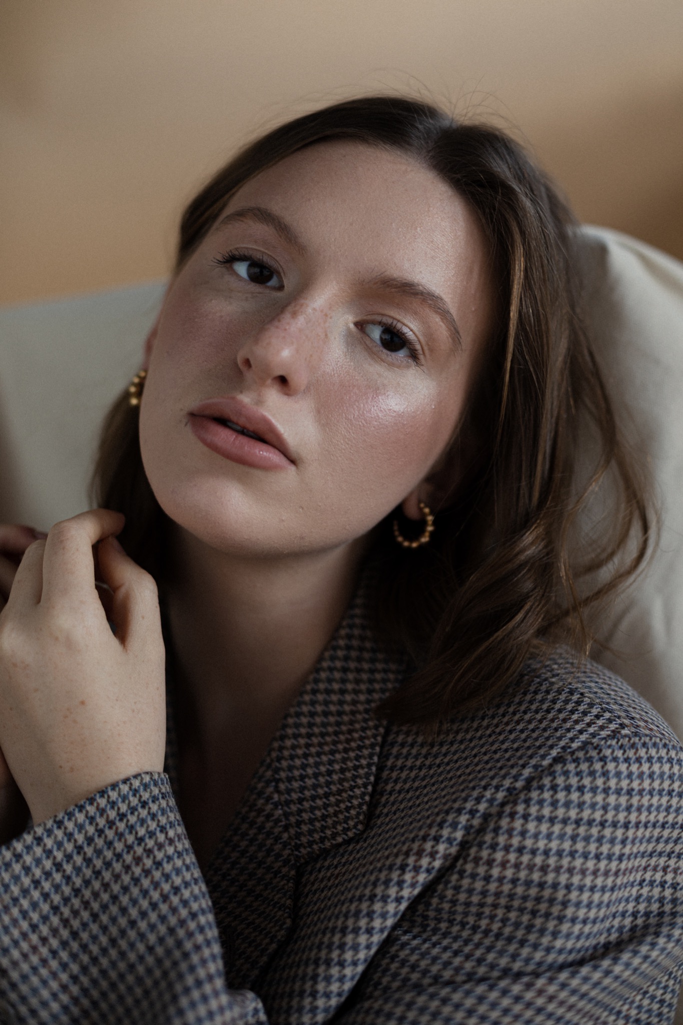 Portret kobiety z delikatnym makijażem, w tweedowej marynarce, pozującej w subtelnym oświetleniu, z widocznymi złotymi kolczykami.