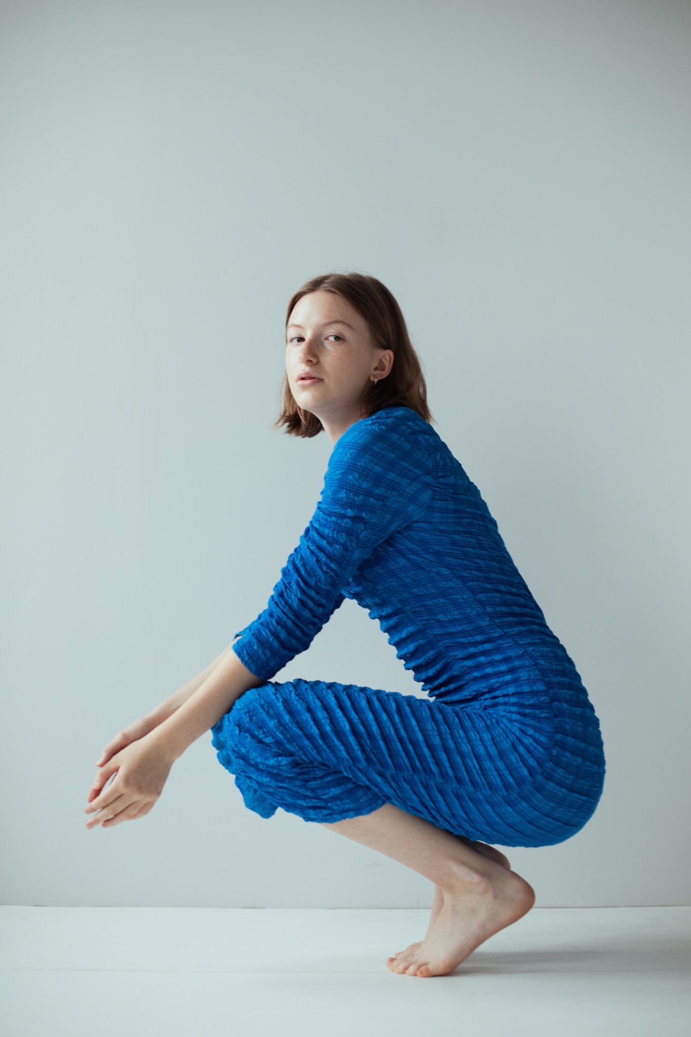 Młoda kobieta w niebieskiej plisowanej sukience kuca na białym tle, spoglądając w obiektyw z delikatnym uśmiechem.