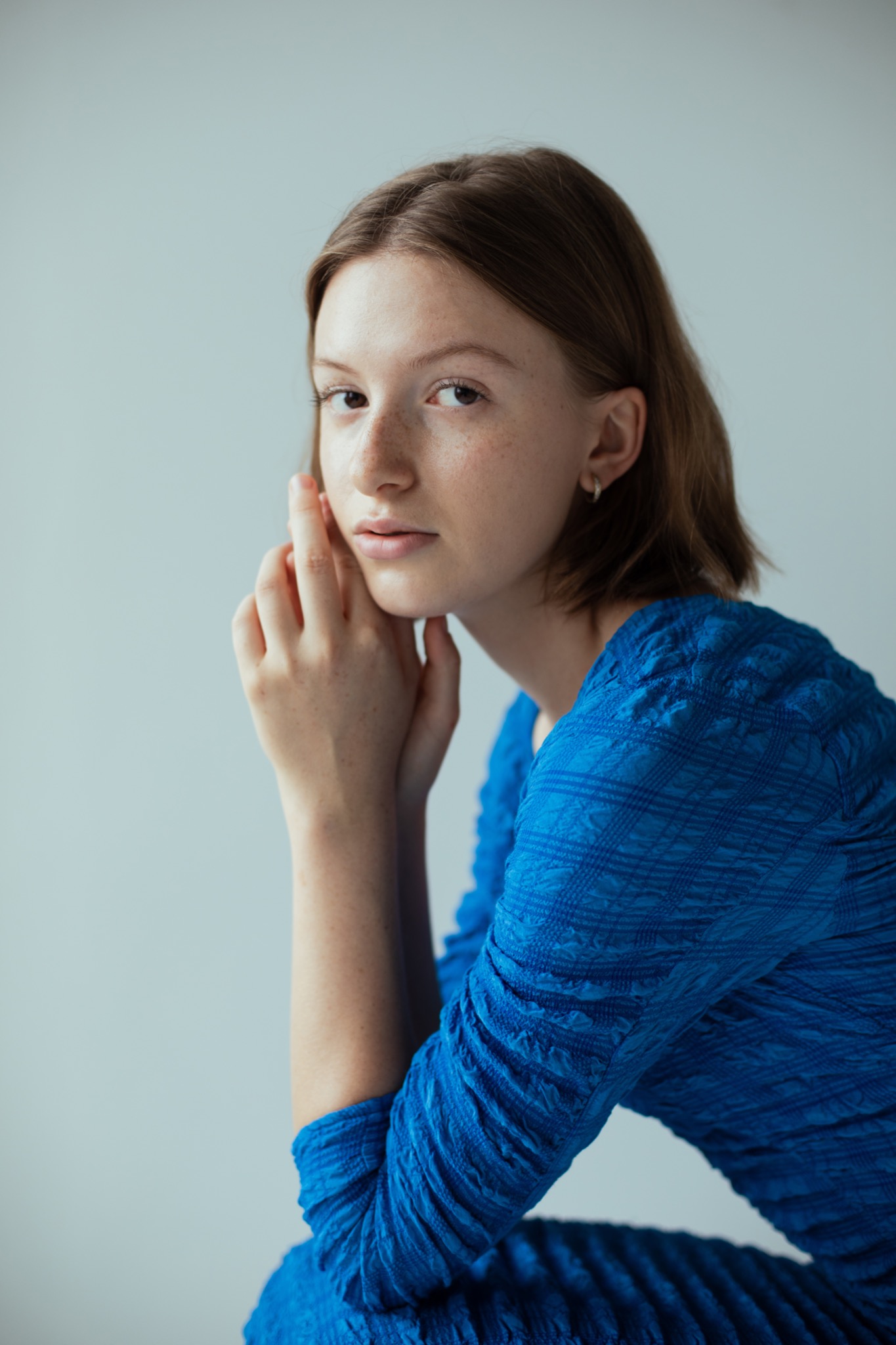 Portret młodej kobiety z piegami, w niebieskiej sukience o teksturowanej fakturze, opierającej dłonie o twarz na neutralnym tle.