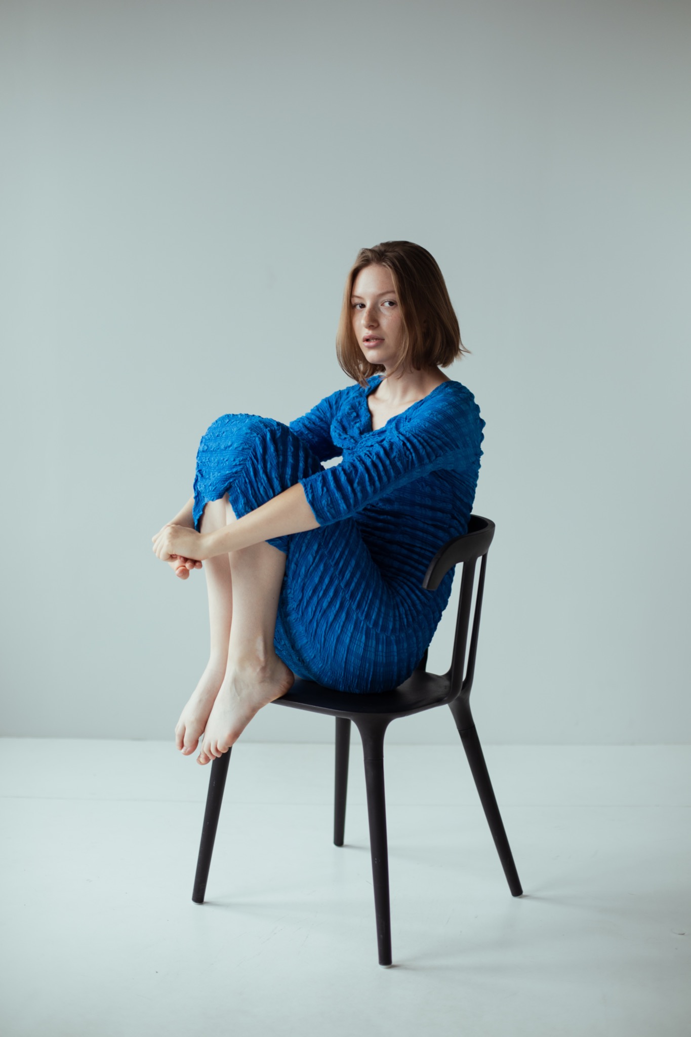 Młoda kobieta w niebieskiej, teksturowanej sukience siedzi na czarnym krześle, obejmując kolana i patrząc w obiektyw na tle jasnoszarego studia.