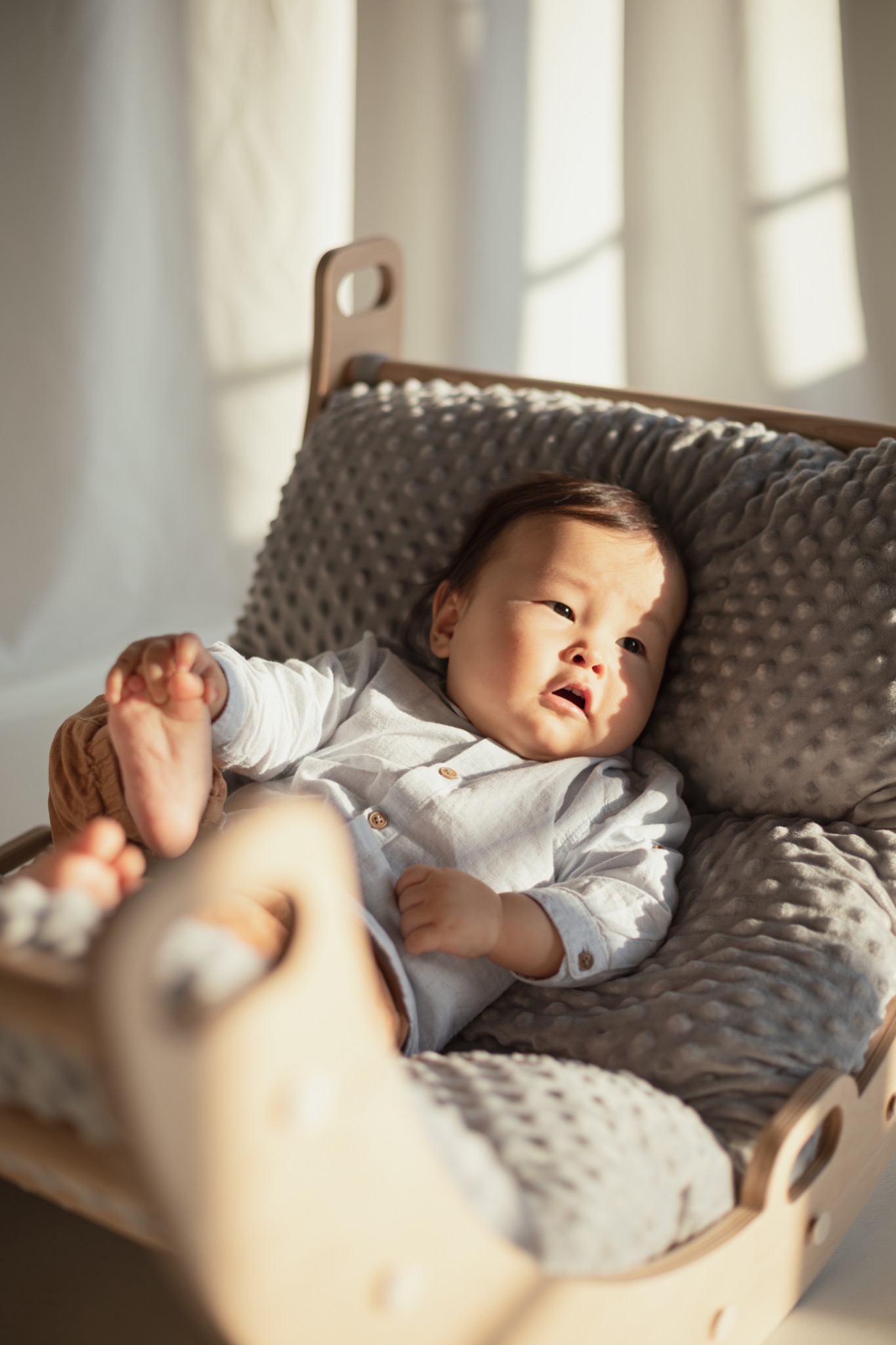 Mały chłopiec w jasnej koszuli leży w drewnianym bujaku z szarą, pluszową poduszką, z promieniami słońca padającymi na jego twarz i tło.