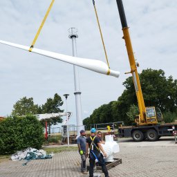 OZE INSTAL - Montaż łopaty turbiny wiatrowej za pomocą dźwigu; widoczni pracownicy w kaskach i uprzężach, w tle wieża turbiny i zieleń.