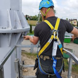 OZE INSTAL - Pracownik w kasku i uprzęży, z tyłu, dokręca śruby na metalowej konstrukcji turbiny wiatrowej, w tle zabudowania mieszkalne i turbina wiatrowa.
