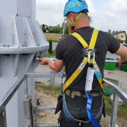 Pracownik w kasku i uprzęży, z tyłu, dokręca śruby na metalowej konstrukcji turbiny wiatrowej, w tle zabudowania mieszkalne i turbina wiatrowa.