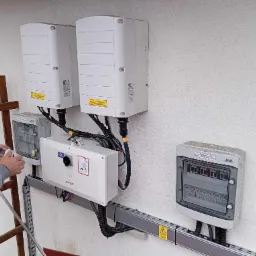 Instalacja fotowoltaiczna: monter czyści obudowę inwertera SolarEdge na zewnątrz budynku, widoczne skrzynki rozdzielcze i drabina.