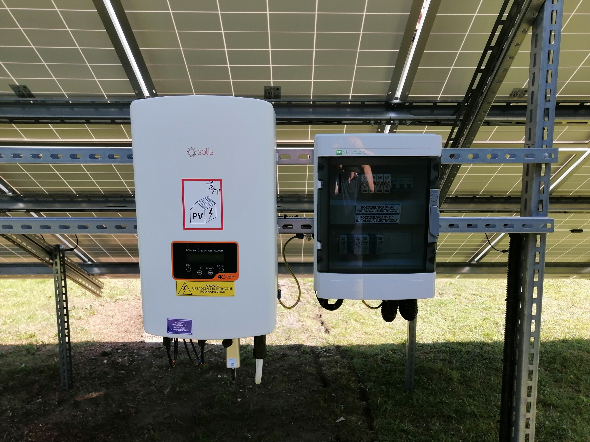 Inwerter marki Solis wraz z rozdzielnicą elektryczną przy panelach słonecznych zamontowanych na konstrukcji wsporczej na zewnątrz.