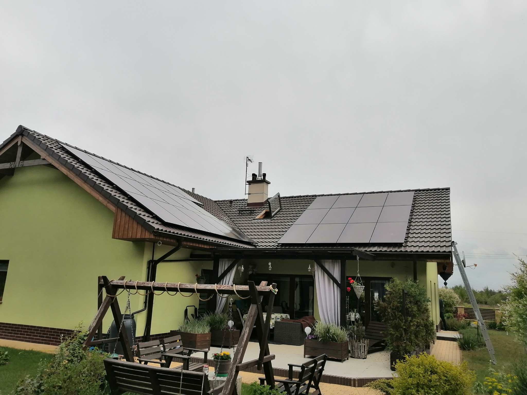 Panele słoneczne zamontowane na dachu domu jednorodzinnego z zieloną elewacją, widok z poziomu ogrodu z drewnianą huśtawką.