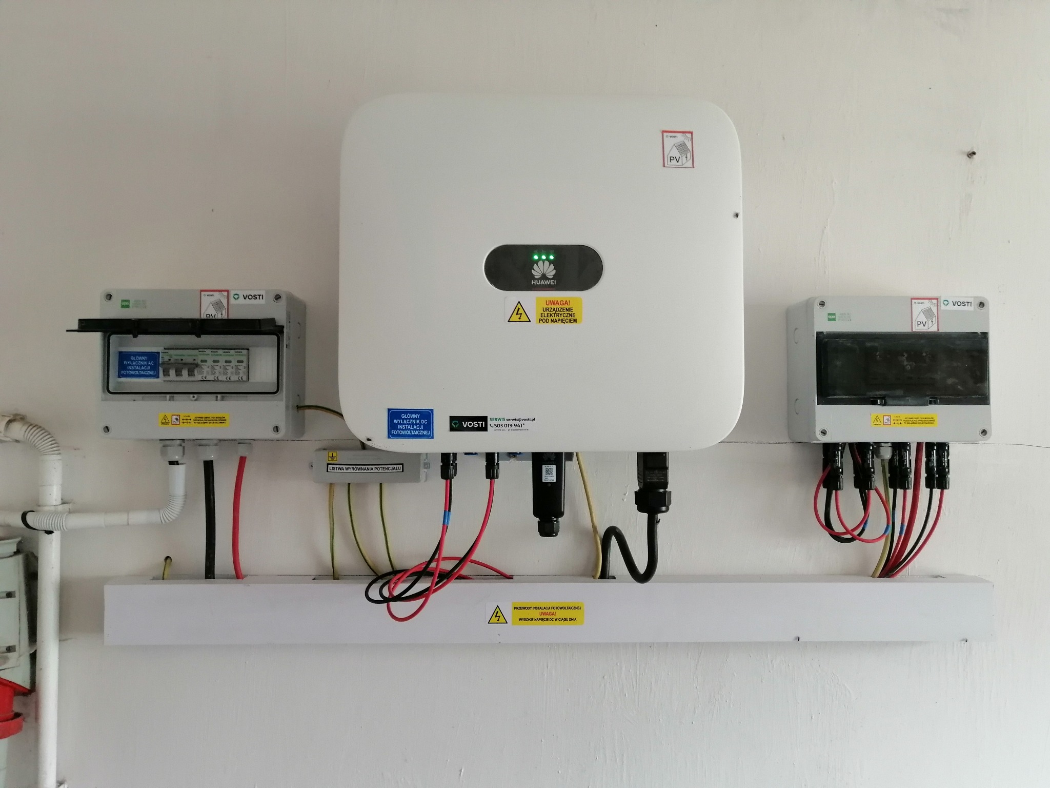 Biały inwerter Huawei z zielonymi diodami, zamontowany na ścianie pomiędzy dwoma szarymi skrzynkami z bezpiecznikami. Pod spodem listwa z kablami. Widoczne naklejki ostrzegawcze i logo Vosti.