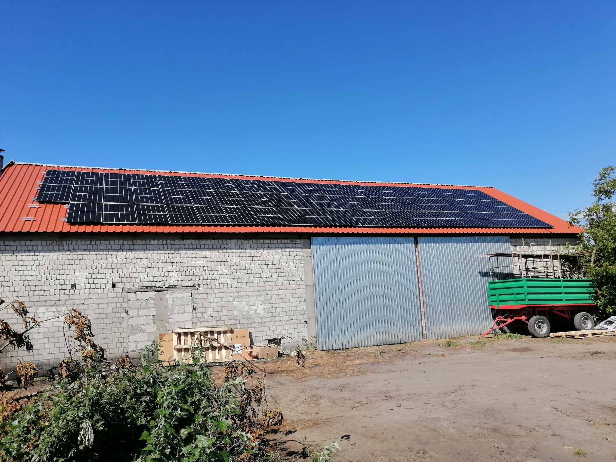 Duży budynek gospodarczy z czerwonym dachem, na którym zamontowano panele słoneczne. Obok budynku stoi zielona przyczepa rolnicza.