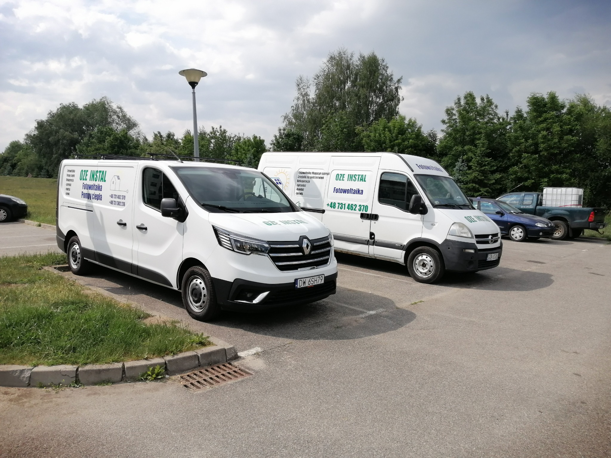 Dwa białe samochody dostawcze firmy OZE Instal z logo i numerem telefonu zaparkowane na parkingu, jeden z nich z widoczną tablicą rejestracyjną.