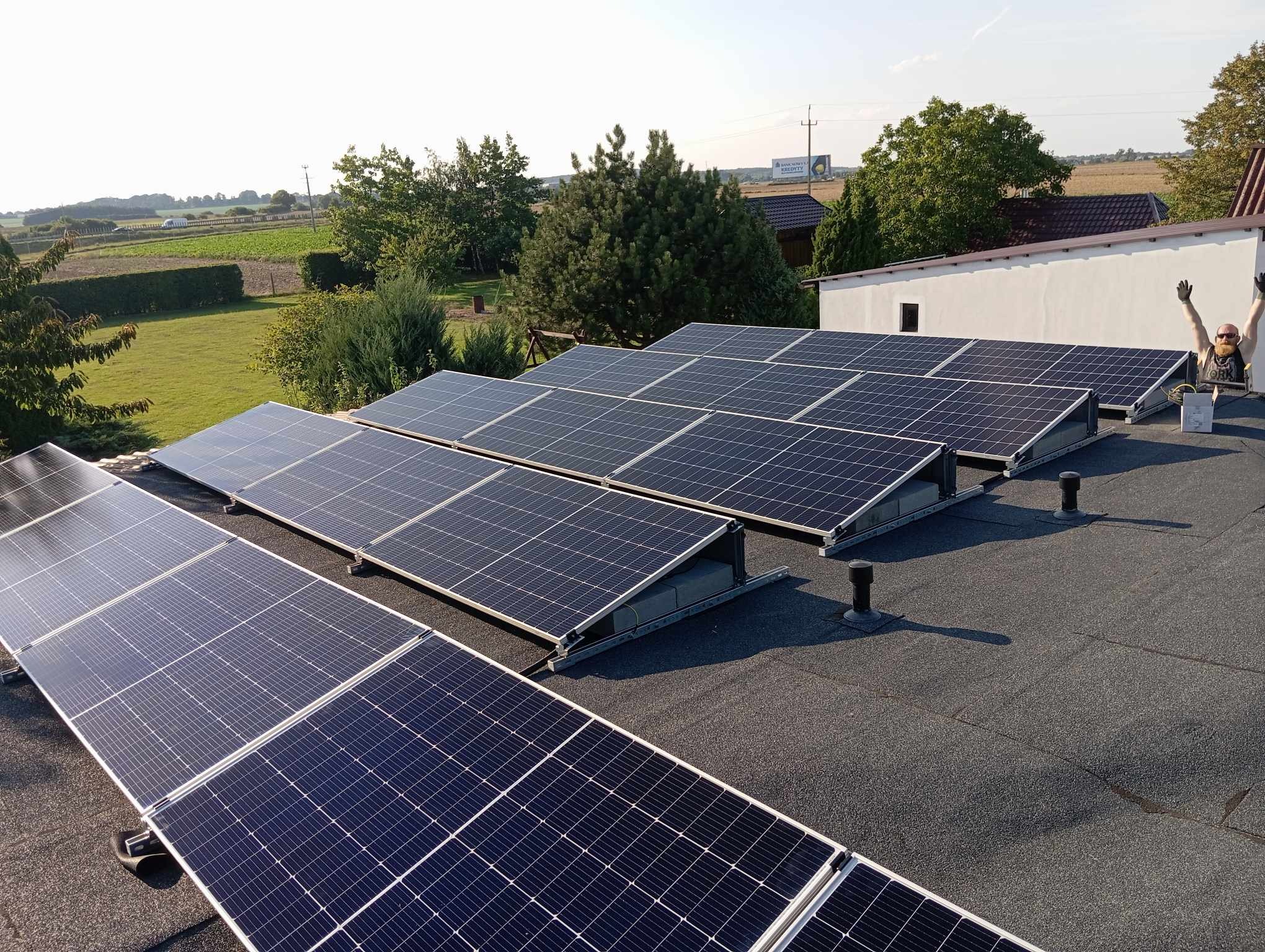 Widok z góry na instalację paneli słonecznych na dachu pokrytym papą, z widocznym instalatorem z podniesionymi rękami w rękawicach, oraz krajobrazem wiejskim w tle.