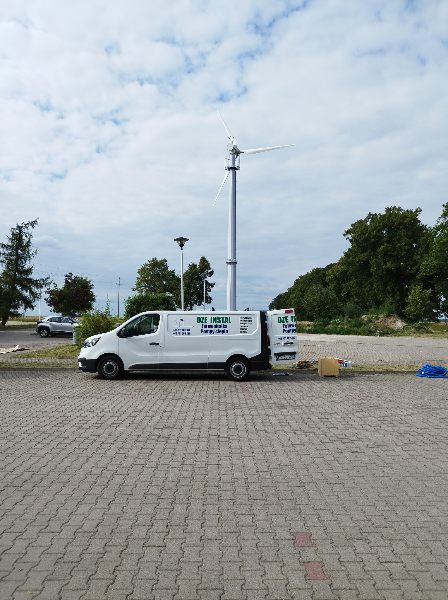 Biały samochód dostawczy z logo firmy OZE INSTAL na tle turbiny wiatrowej i pochmurnego nieba. Na samochodzie widoczne informacje o fotowoltaice i pompach ciepła.