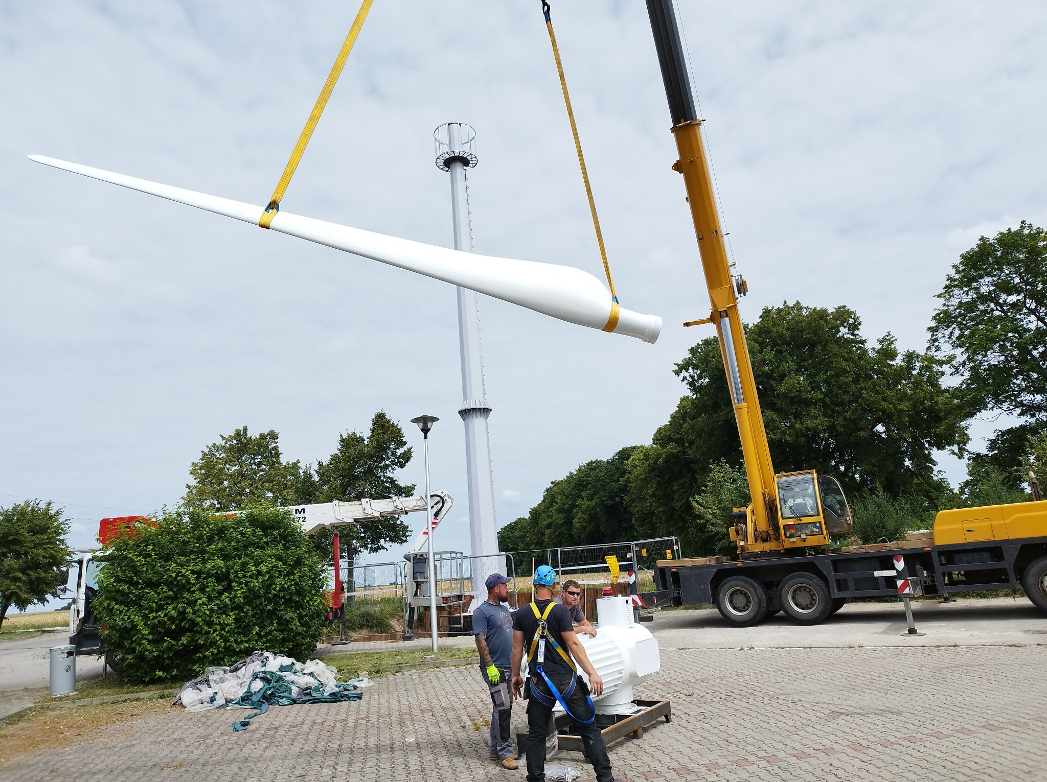 Montaż łopaty turbiny wiatrowej za pomocą dźwigu; widoczni pracownicy w kaskach i uprzężach, w tle wieża turbiny i zieleń.