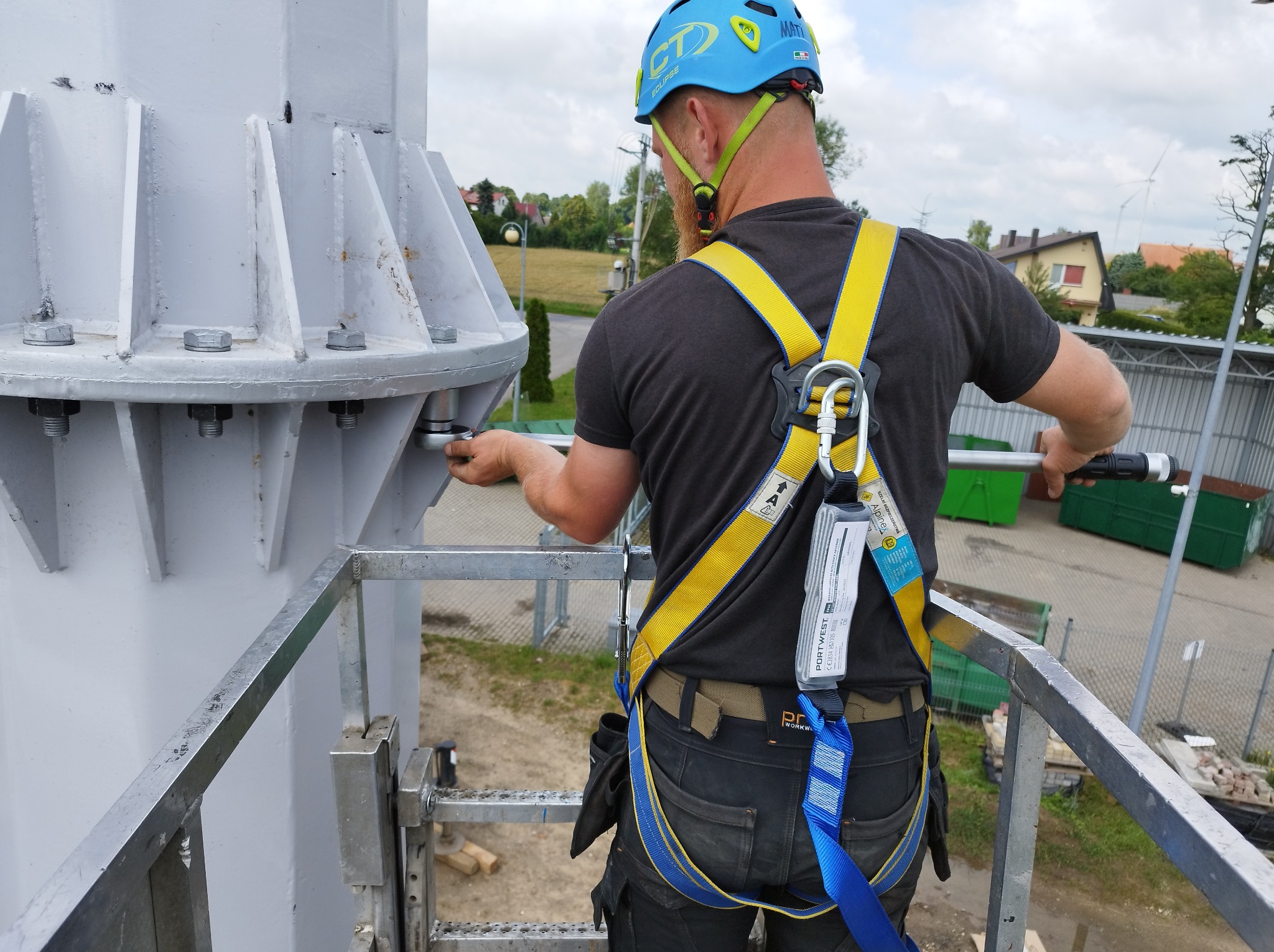 Pracownik w kasku i uprzęży, z tyłu, dokręca śruby na metalowej konstrukcji turbiny wiatrowej, w tle zabudowania mieszkalne i turbina wiatrowa.