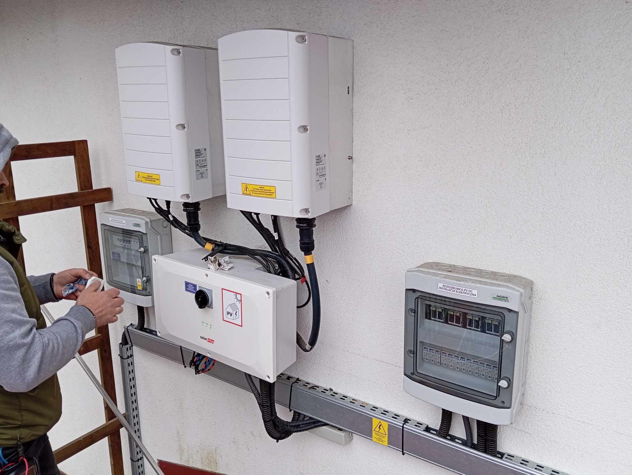 Instalacja fotowoltaiczna: monter czyści obudowę inwertera SolarEdge na zewnątrz budynku, widoczne skrzynki rozdzielcze i drabina.