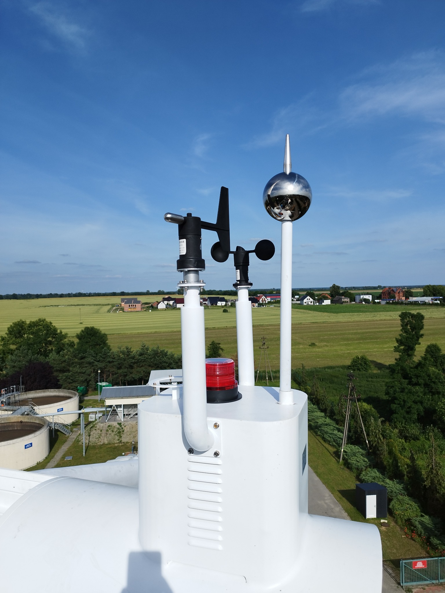 Biała instalacja z metalową kulą i wiatromierzem na dachu, z widokiem na pola i budynki w tle pod błękitnym niebem.