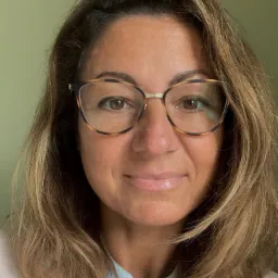 Portret uśmiechniętej kobiety w okularach o wzorze panterki, z brązowymi włosami z pasemkami, na jednolitym, jasnym tle.