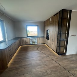 DAREKMEBLE Dariusz Głaz - Nowoczesna kuchnia z drewnianymi frontami, podświetleniem LED i czarnymi blatami. Widok na okno z zielenią w tle. Częściowo widoczne gniazdka elektryczne na ścianie.