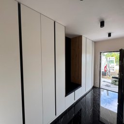 DAREKMEBLE Dariusz Głaz - Biała szafa wnękowa z siedziskiem z ciemnego drewna i czarną tapicerką, na tle błyszczącej, czarnej podłogi. Widok na korytarz i otwarte drzwi wejściowe.