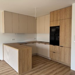 DAREKMEBLE Dariusz Głaz - Nowoczesna kuchnia w kształcie litery U z drewnianymi i beżowymi frontami, czarnym piekarnikiem i płytą indukcyjną. Widoczne detale wykończenia i oświetlenie.