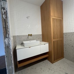 DAREKMEBLE Dariusz Głaz - Nowoczesna łazienka z białą umywalką nablatową, szafką z drewnianym akcentem i wysoką szafą. Ściany wyłożone białymi płytkami w jodełkę i szarym lastryko.