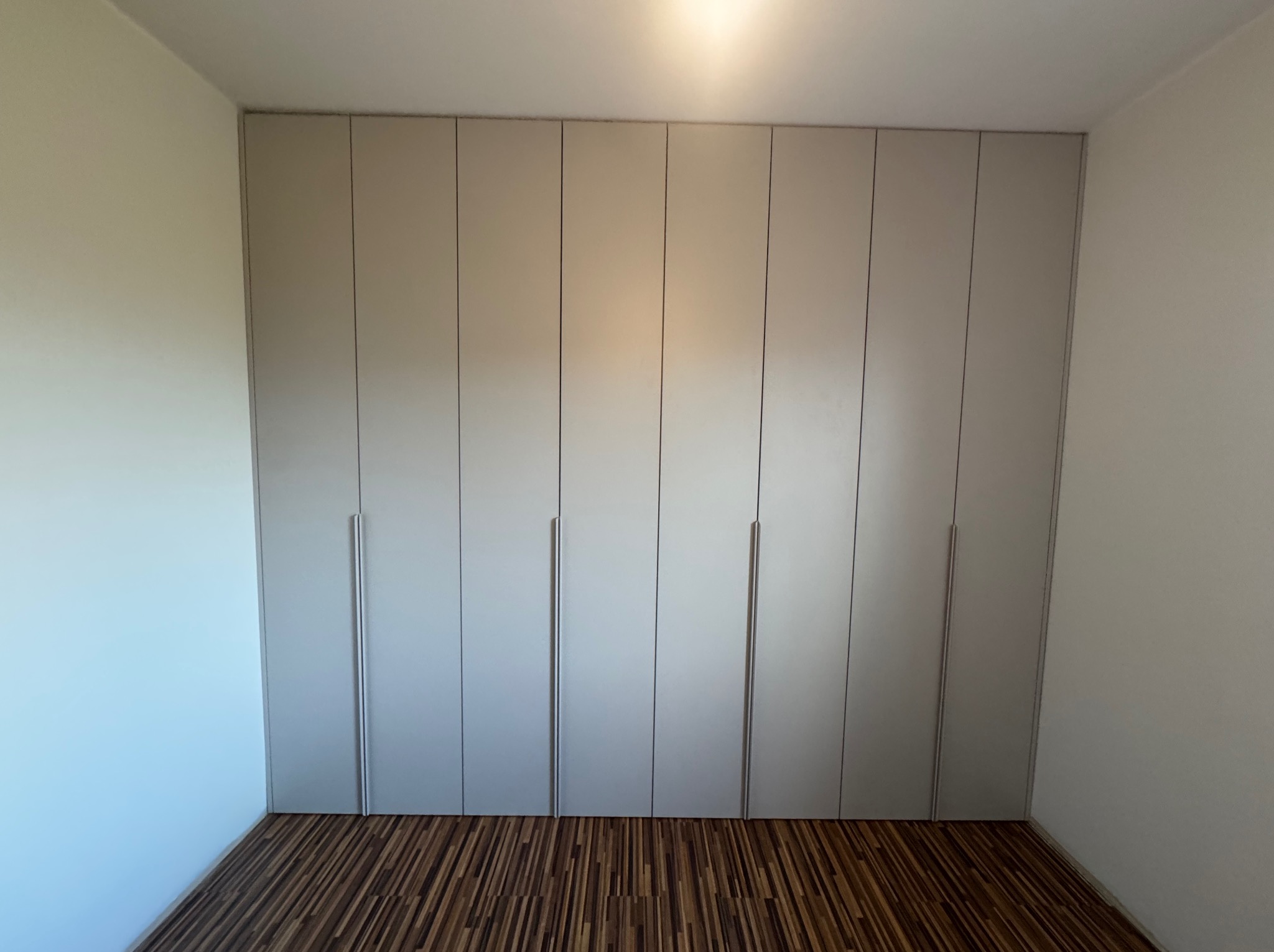 Jasnoszara szafa wnękowa z długimi, pionowymi uchwytami, idealnie dopasowana do wnętrza z drewnianą podłogą. Minimalistyczny design.