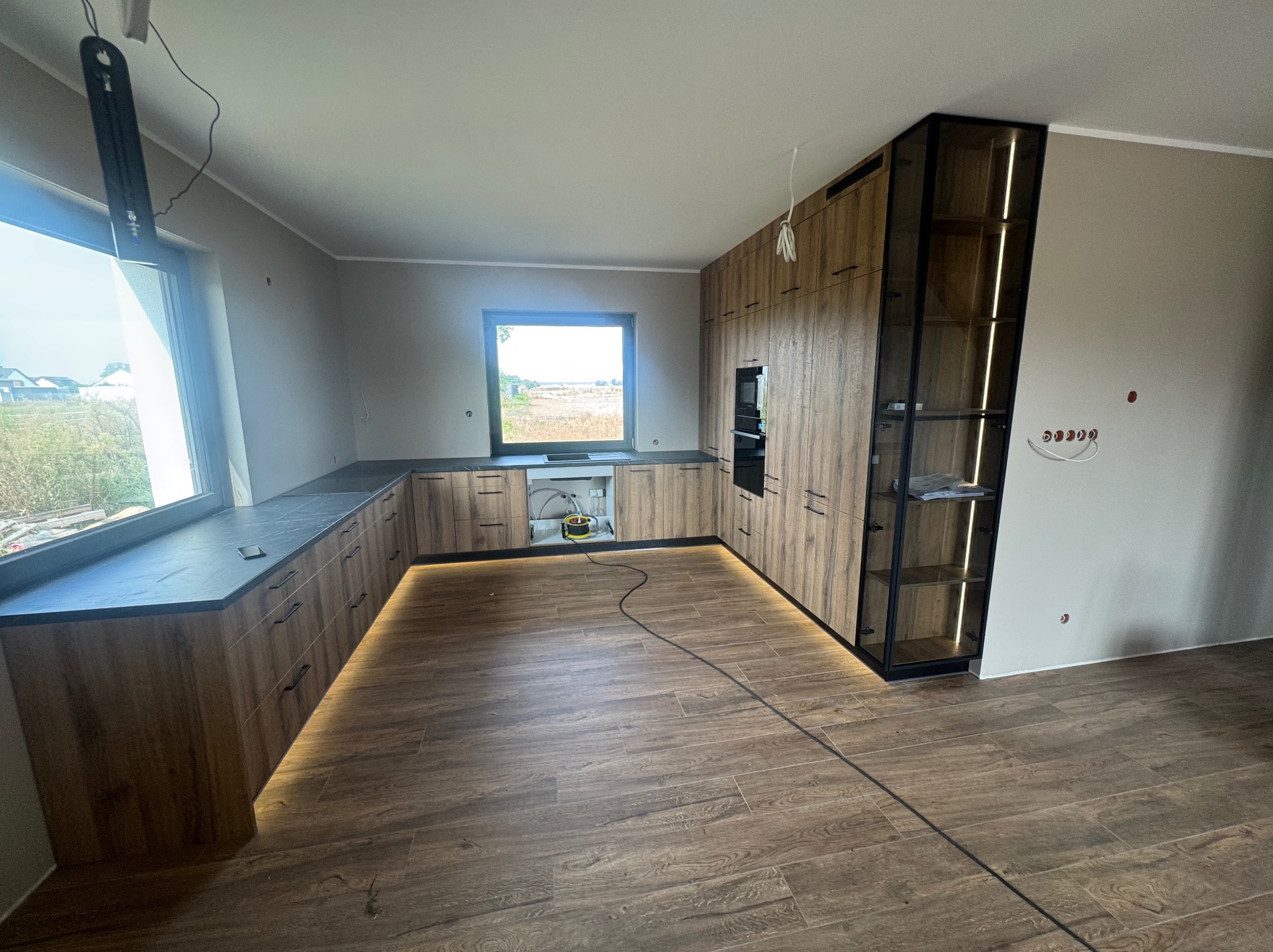 Nowoczesna kuchnia z drewnianymi frontami, podświetleniem LED i czarnymi blatami. Widok na okno z zielenią w tle. Częściowo widoczne gniazdka elektryczne na ścianie.