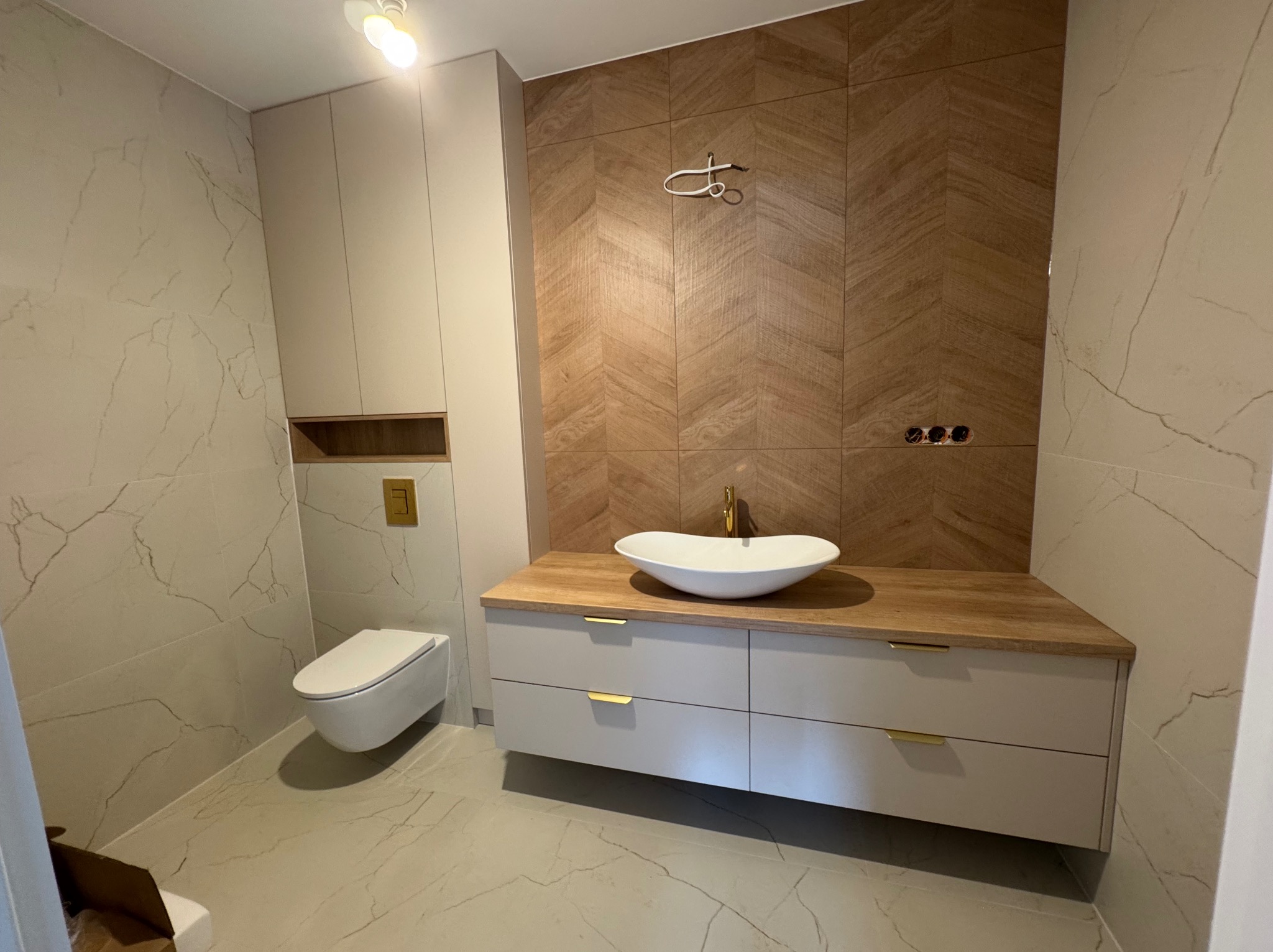 Nowoczesna łazienka z drewnianą ścianą w jodełkę, umywalką nablatową i szafką z szufladami. Uwagę zwraca wisząca toaleta i marmurowe płytki.